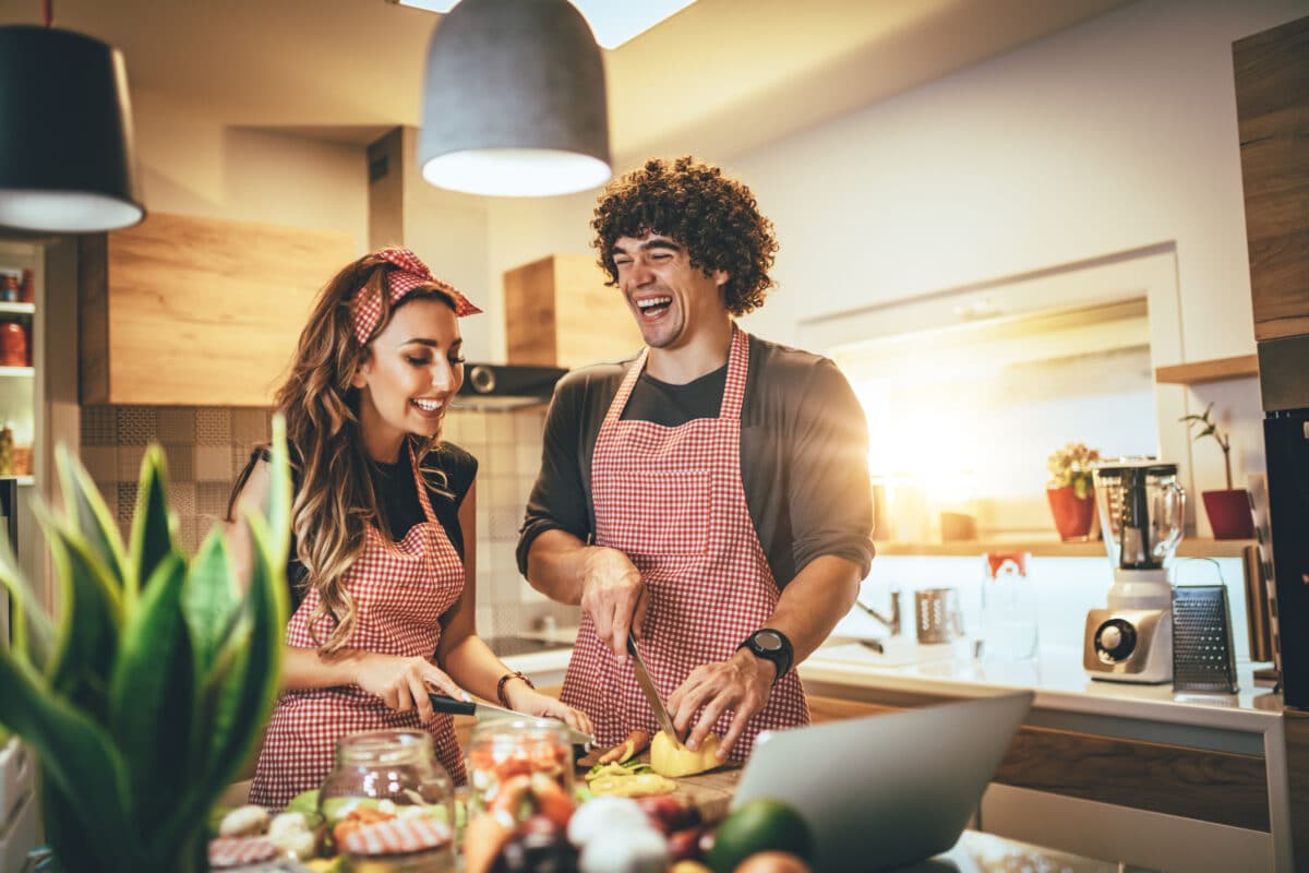 A couple laughing and cooking together in the kitchen while cutting vegetables.