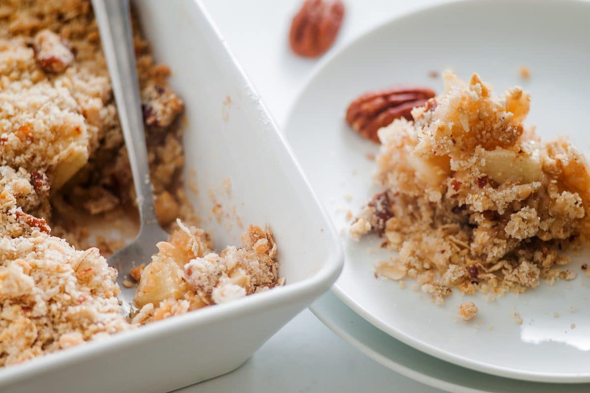 A corner view of the baking dish with a spoon in it and a white dish to the right with a piece of apple pecan crisp on it