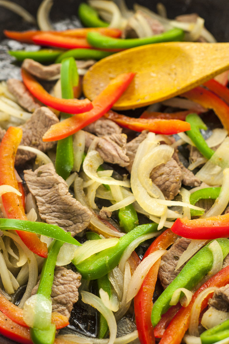 steak, peppers, and onions being cooked in a pan with a wooden spoon