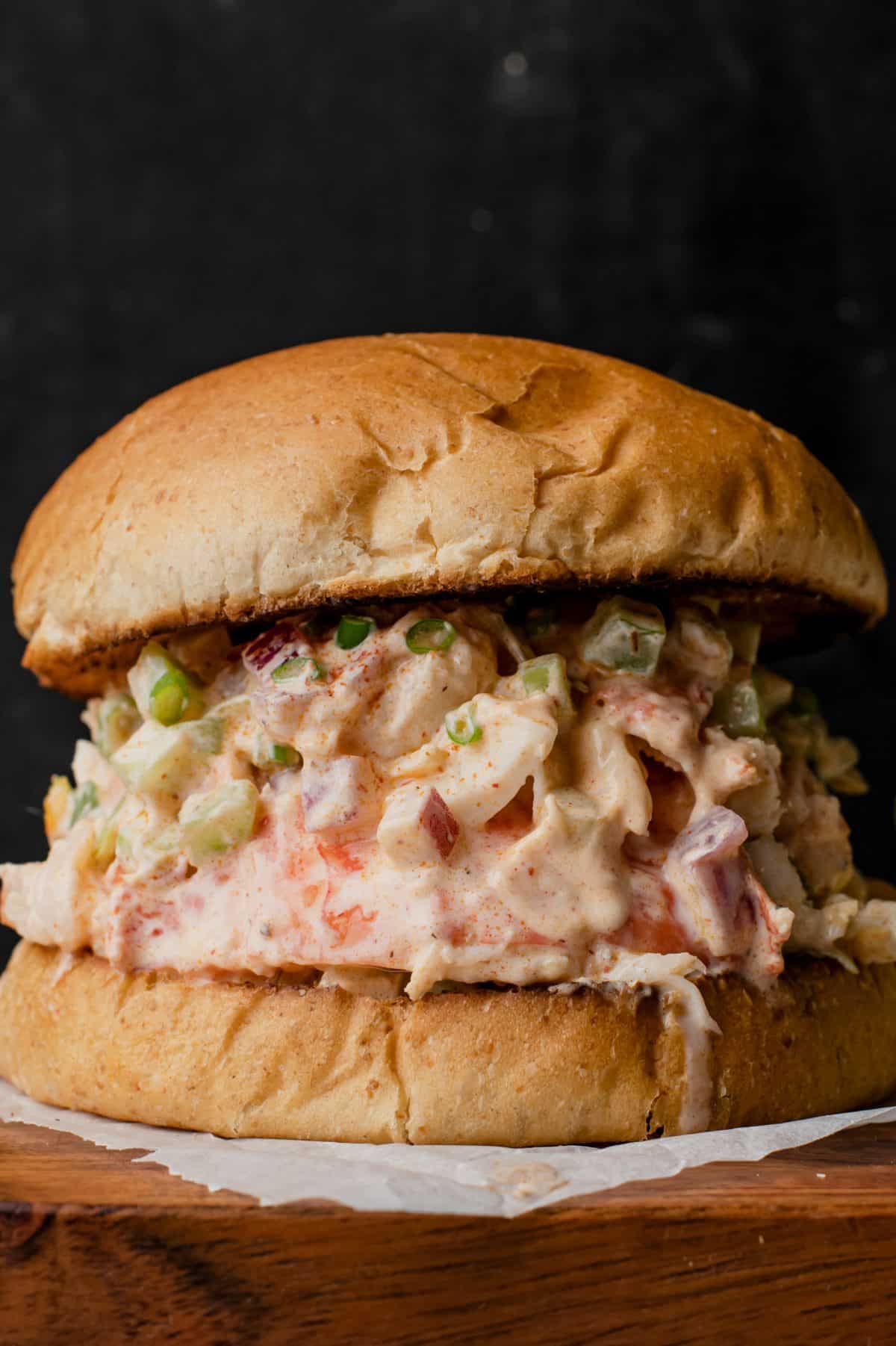 tall image of lobster salad between slices of bun on black background and wooden cutting board