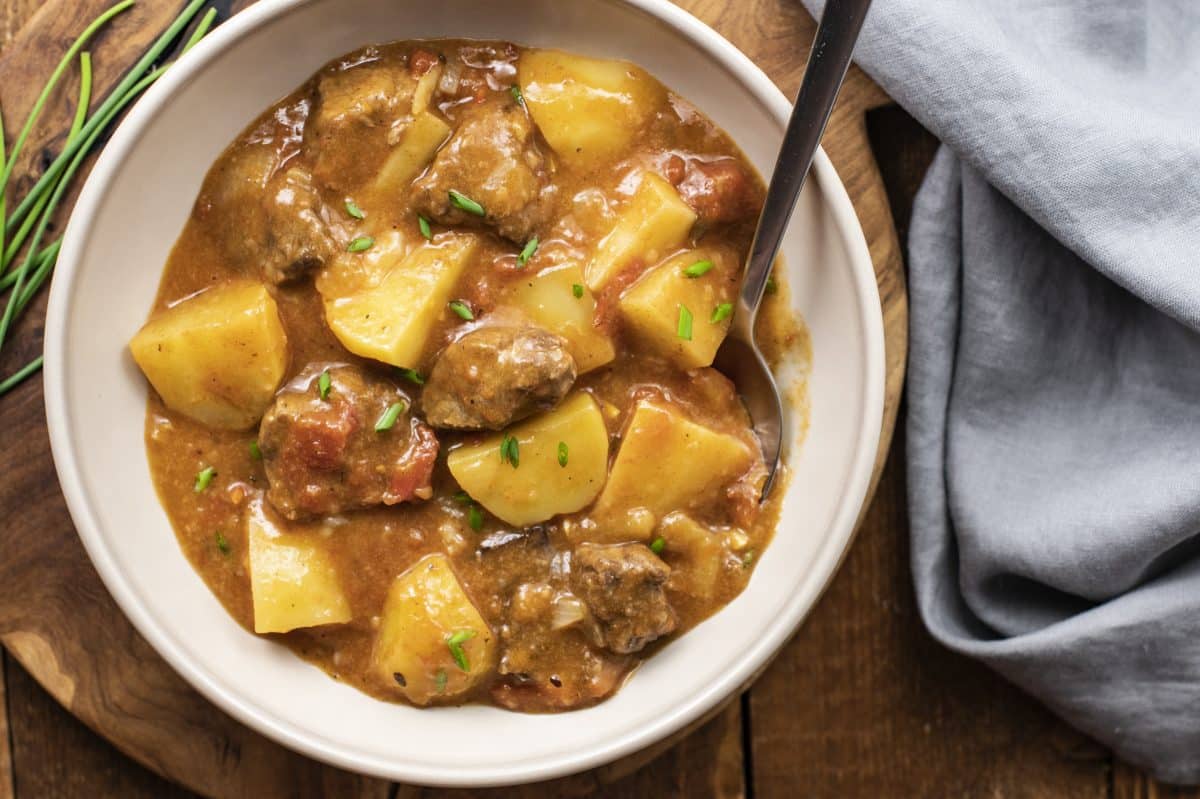 beef and potato stew in a white bowl with a spoon and a greyish blue napkin on the side