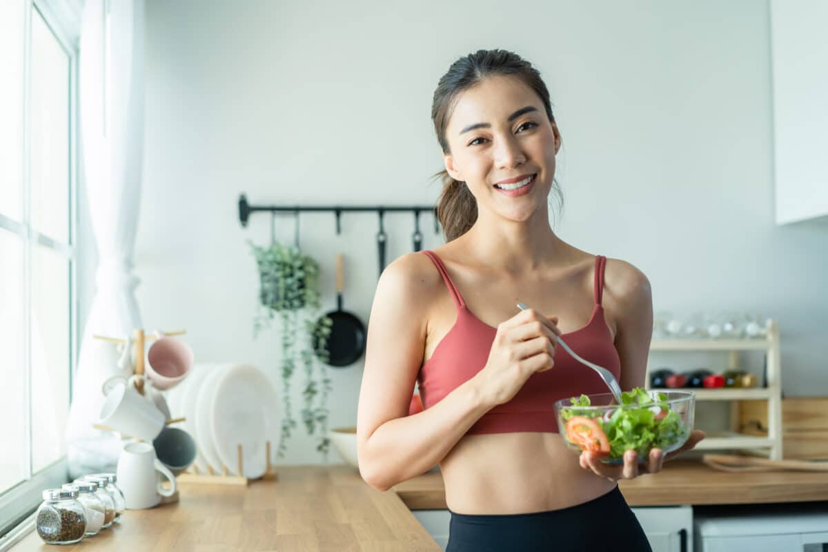 Asian woman eating a salad in workout clothes in the kitchen.