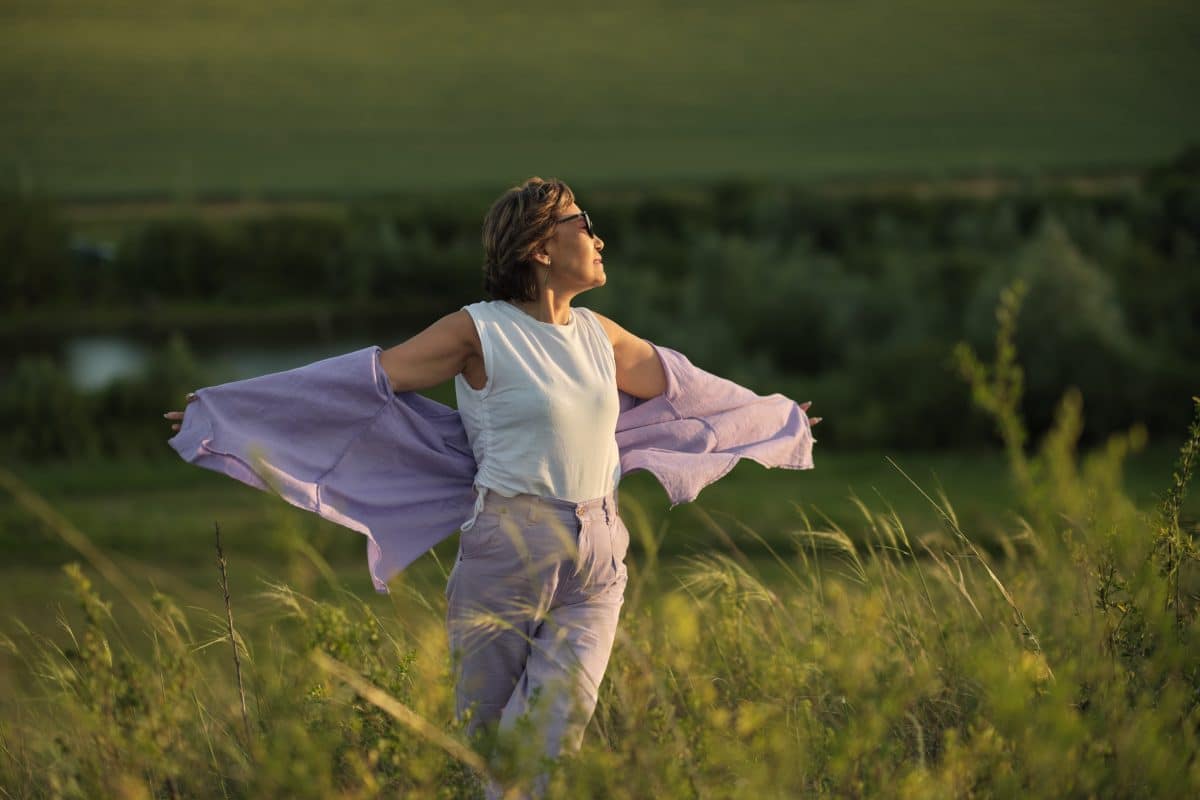 Middle-aged woman with arms out in a field during sunset.