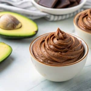 Homemade chocolate mousse recipe in a white bowl with avocado in the background.