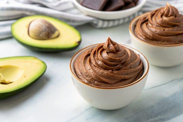 Homemade chocolate mousse recipe in a white bowl with avocado in the background.