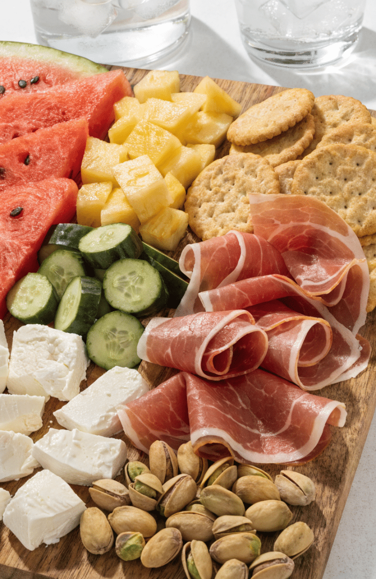 Watermelon, pineapple, cucumber, feta, crackers, and prosciutto on a charcuterie board.